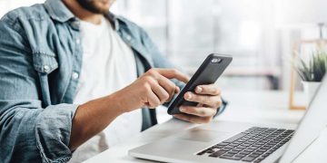 Man scrolling on phone next to laptop
